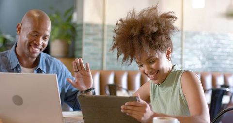 Young professionals collaborating in cozy cafe: smiling woman holding tablet, man waving