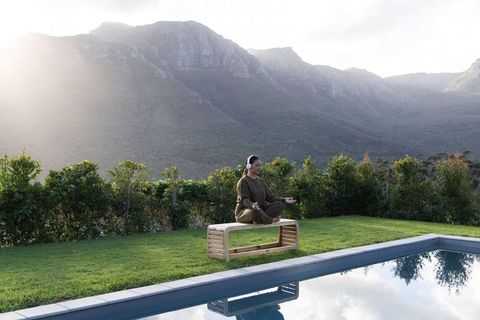 Asian woman meditating on low bench by pool with headphones amid mountain views wellness