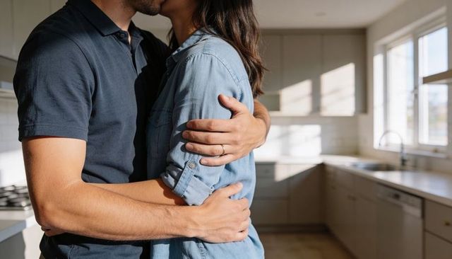 Couple kissing and hugging in sunlit modern kitchen showing wedding ring