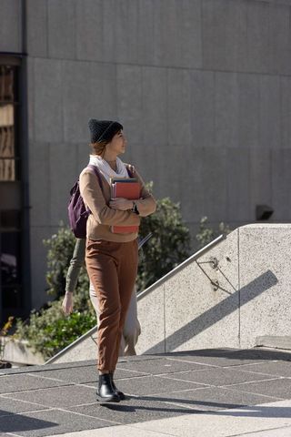 Asian student standing on urban campus plaza holding notebooks and wearing knit beanie