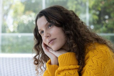 Contemplative young woman wearing mustard sweater gazing out sunlit window with soft bokeh