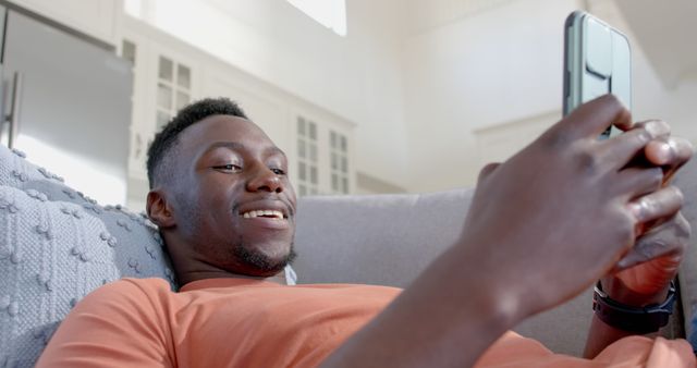 Man lying comfortably on a sofa, smiling while looking at a smartphone. Could be used for themes related to home life, relaxation, using technology, or personal leisure time. Suitable for advertising lifestyle apps, home decor items, or technology products.