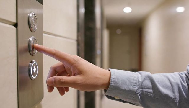 Right hand pressing elevator button with illuminated down arrow in modern building hallway