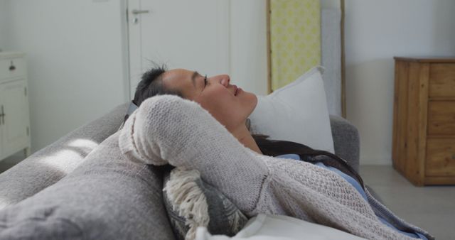 Woman sitting on a grey couch in a home interior, wearing a light-colored casual sweater. She appears relaxed with her head resting on the back of the couch, suggesting tranquility and comfort. Ideal for use in lifestyle magazines, relaxation and mental health articles, or advertisements depicting comfortable home living or leisure time.