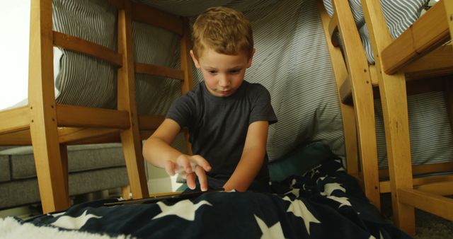 This image shows a young boy engaging in imaginative play inside a home-made fort. He is using a tablet while being surrounded by blanket and framed by furniture. Perfect for uses related to childhood activities, technology use among children, indoor play, creativity, and family life.