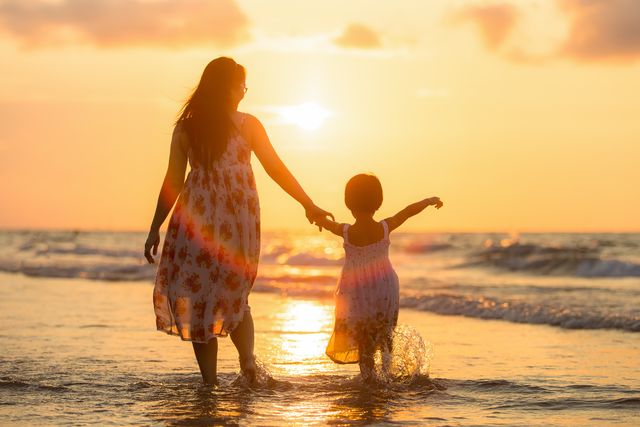 This image shows a mother and her child walking hand in hand along a beach at sunset. The sun casts a warm glow over the scene creating a peaceful and serene atmosphere. Ideal for themes of family, love, summertime, vacations, and peaceful retreats.