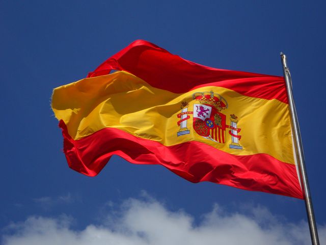 Spanish flag prominently waving against a clear blue sky, representing national pride and patriotism. Ideal for use in educational materials about Spain, travel brochures, cultural studies, government presentations, and international relations content.