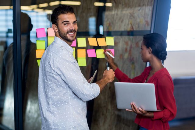 Colleagues collaborating in an office, using sticky notes on a glass wall for brainstorming and planning. Ideal for business, teamwork, and productivity concepts. Useful for illustrating collaborative work environments, strategic planning sessions, and creative meetings.