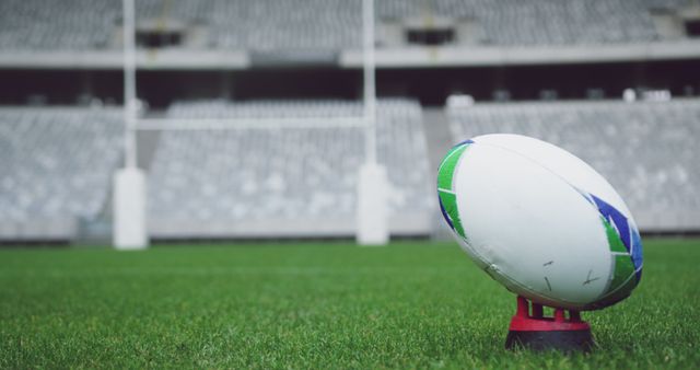 Close-up of a rugby ball placed on a kick-off tee on a green field in an empty stadium. Ideal for use in sports promotions, advertisements for rugby equipment, articles discussing rugby games or techniques, and visual content for fan engagement.