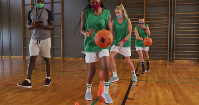 Female basketball players are practicing dribbling skills while wearing face masks in a gym with their coach. This image can be used to illustrate themes of women's sports, health and safety during the pandemic, team training, athleticism, and indoor fitness activities. It is suitable for articles, advertisements, and promotional materials related to sports education, safety protocols, and women's empowerment in sports.