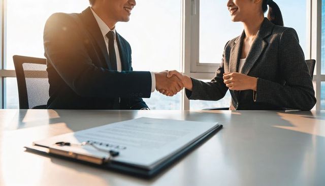 Business partners shaking hands over signed contract in sunlit office conference room