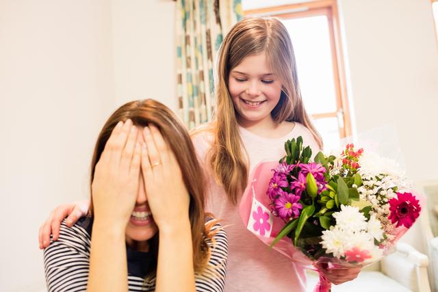 Mother covering her eyes while receiving gift from daughter in living room