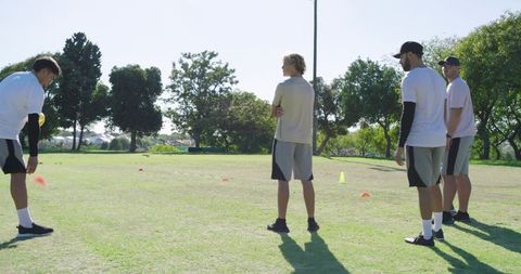 Diverse male team training soccer drills on sunny park field with yellow ball and cones
