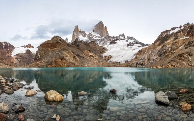Panoramic view showcasing the Fitz Roy mountains in Argentina, reflecting over a calm lake surrounded by a rocky shore. The snow-covered peaks and the glacier create a dramatic and serene landscape. Ideal for use in travel blogs, adventure promotions, nature documentaries, and outdoor activity advertisements.