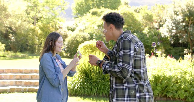 Young couple enjoying blowing bubbles together in a sunny park. Ideal for content depicting outdoor activities, playful interactions, and young adult lifestyle. Perfect for use in social media posts, happiness and leisure themed advertisements, or articles about summer fun.