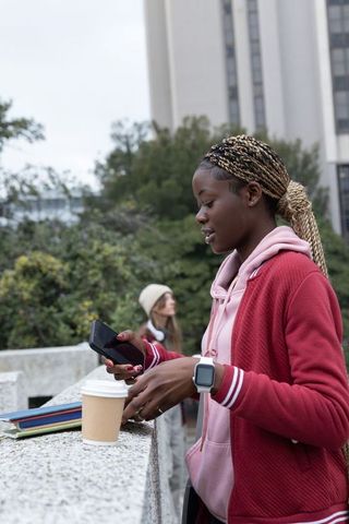 Teenage woman checking phone and steadying coffee at campus railing