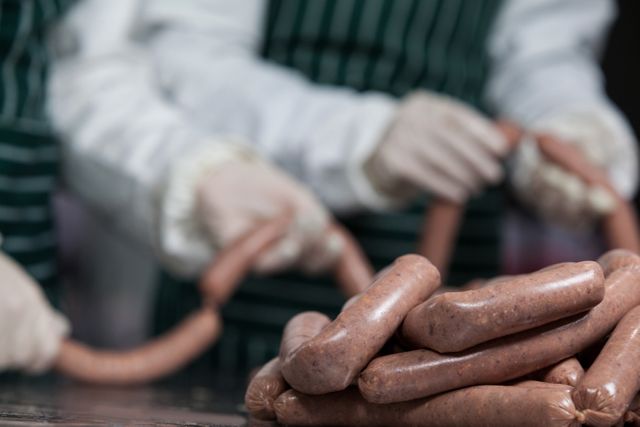 Butchers wearing gloves are processing and preparing sausages in a meat factory. This image captures the meticulous handling and hygiene standards in food preparation in the meat industry. Useful for articles or promotions related to food safety, meat production, professional culinary skills, and factory operations.