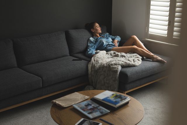 Woman lounging on a dark grey sofa in a modern living room. She is wearing casual clothes and appears relaxed. A soft blanket is draped over the sofa. Ideal for use in lifestyle blogs, home decor websites, or advertisements promoting relaxation and comfort in home settings.