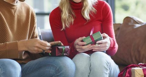 Couple exchanging ribboned gifts on sofa, woman in red turtleneck, cozy holiday moment