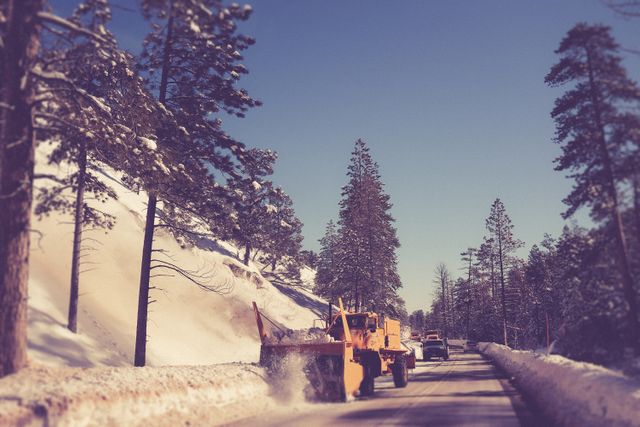 Snow plow in action on a mountain road covered in snow. Snow-covered trees are visible alongside the road under a clear blue sky. Ideal for illustrating winter road services, transportation in winter weather, and cold climate maintenance tasks.