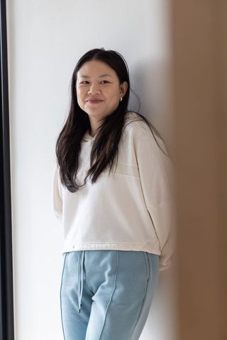 Asian woman leaning on white wall in soft natural light wearing cream hoodie and blue sweatpants