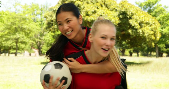 Two young female soccer players wear matching red and black uniforms, smiling and embracing with a soccer ball in hand, on a sunny field. Perfect for promoting team sports, friendship, and youth recreational activities.