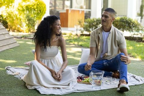 Young couple laughing and sitting on picnic blanket in backyard garden with fresh fruit