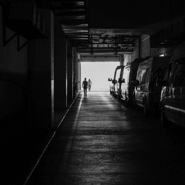 Person walking alone in a dimly lit underground parking garage with light at the end of the corridor. Several parked vehicles seen along the walls. Capturing urban solitude and industrial atmosphere. Used for illustrating urban settings, solitude themes, and industrial backgrounds.