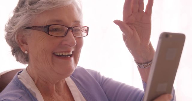 Elderly woman with glasses smiling and waving at a smartphone while making a video call. Great for illustrating concepts about staying connected, elderly people using technology, families communicating, and promoting communication services for seniors.