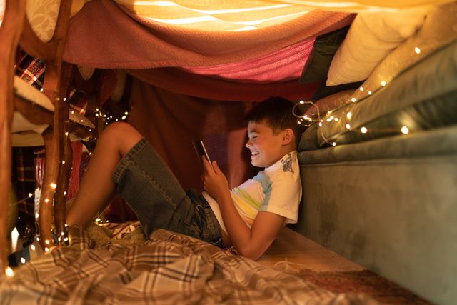 Side view of a caucasian boy sitting on the floor leaning on the couch inside his blanket fort while holding a phone. the fort is lit up with christmas lights