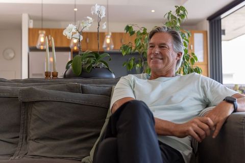 Mature man relaxing on gray sofa smiling while wearing smartwatch in modern open-plan home