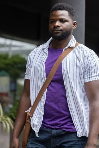 African man standing on urban sidewalk wearing purple t-shirt and brown crossbody bag