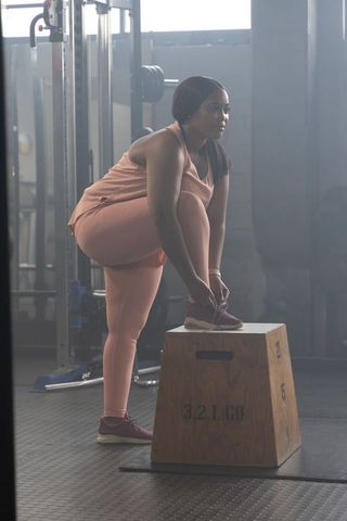 African american woman tying sneaker on plyo box in gym wearing pink activewear