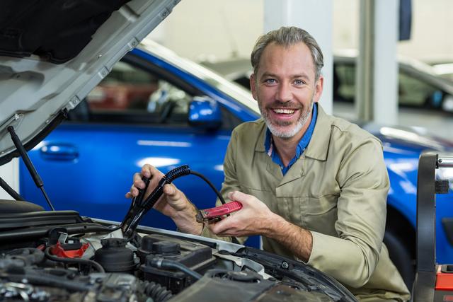 Mechanic in a repair garage attaching jumper cables to a car battery while smiling. Ideal for use in automotive service advertisements, repair shop promotions, and articles about car maintenance and repair services.
