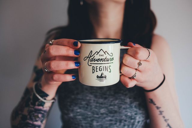 Woman holding a mug with the words 'The Adventure Begins'. Person is wearing a sleeveless top, showcasing rings and forearm tattoos. Ideal for use in personal blogs, motivational webpages, lifestyle magazines, or coffee shop advertisements, emphasizing a sense of beginning a journey or embracing new adventures.