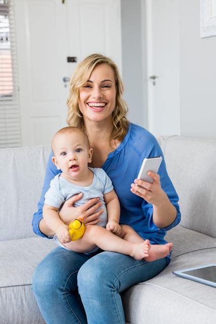 Portrait of mother holding baby boy and using mobile phone in living room at home