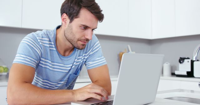 In this stock photo, a man appears frustrated while using his laptop in a clean, modern kitchen. His facial expression indicates displeasure and concern, suggesting he is experiencing issues with his work or technology. The setting includes various kitchen appliances and a sleek countertop, providing a contemporary domestic environment. Ideal for illustrating themes related to remote work challenges, pandemic stress, technological difficulties, work-life balance, and mental fatigue. Suitable for use in corporate presentations, articles on remote working, mental health blogs, or stress management resources.