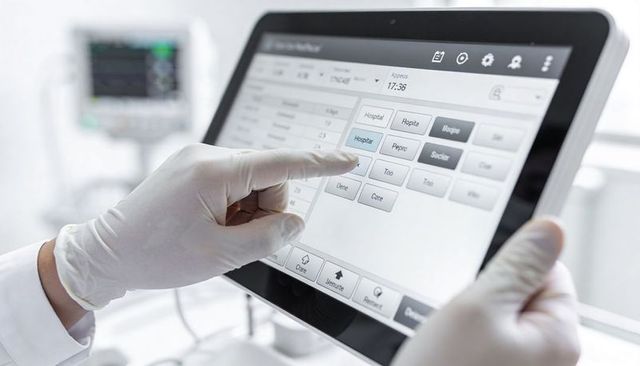 Gloved clinician tapping medical tablet at bedside with vital monitor and touchscreen ui