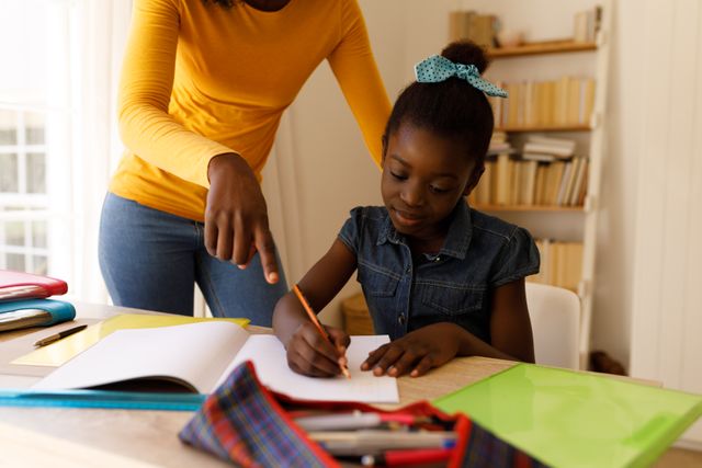 African-American mother assisting her daughter with homework at home. Shelves filled with books in the background suggest a learning environment. Ideal for use in educational materials, parenting blogs, and family support resources.
