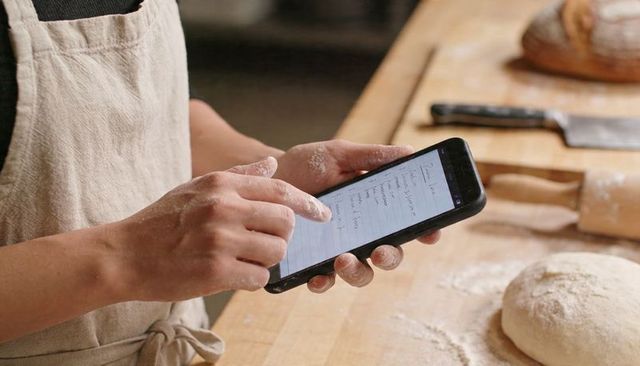 Baker checking digital recipe with floured hands on wooden workbench with dough for artisan bread