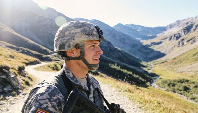 Soldier in digital camo scanning winding mountain trail holding rifle in alpine valley