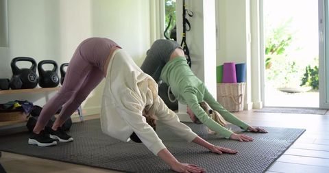 Two women practicing downward dog on mat for home workout with kettlebells and yoga props