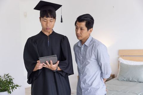 Asian graduate wearing gown and mortarboard checking tablet with father in bedroom