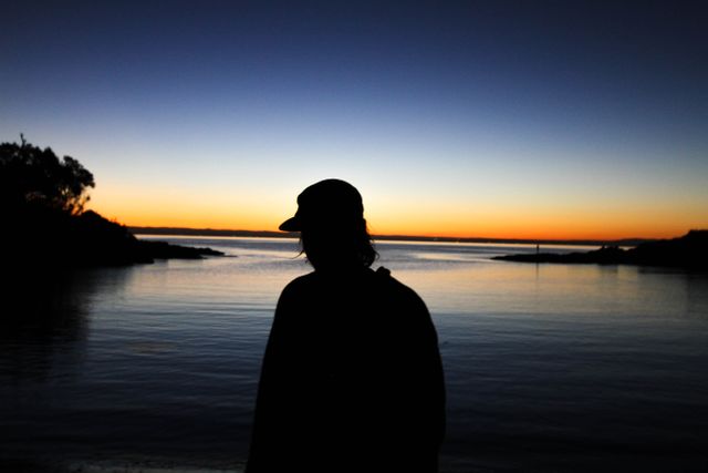 Silhouette of a man standing by the edge of calm water during sunset. Ideal for themes of tranquility, relaxation, evening reflection, and serene nature scenes. Can be used for blog posts, social media banners, inspirational quote backdrops, or serene landscape promotions.