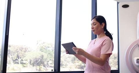 Nurse in Pink Scrubs Checking Tablet Beside MRI Scanner Near Large Window for Radiology