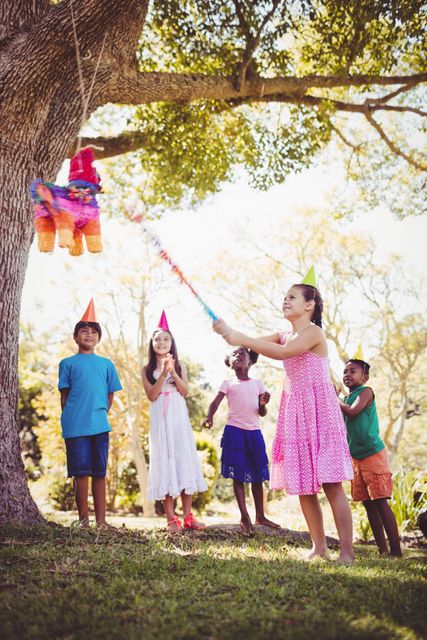 Children enjoying a birthday party in a park, with a girl about to hit a colorful pinata hanging from a tree. Ideal for use in content related to children's parties, outdoor activities, celebrations, and family fun.