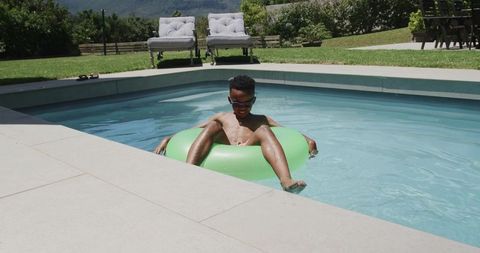 African american teen floating on green inflatable ring in private backyard pool