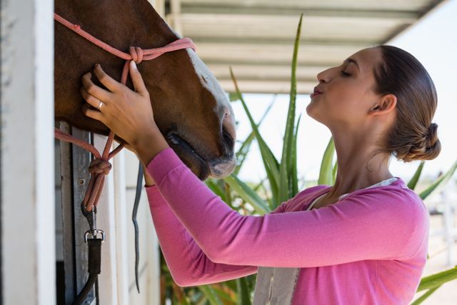Young woman in pink sweater gently touching horse at stable. Ideal for themes related to animal care, equestrian activities, rural lifestyle, and human-animal bonding. Suitable for use in advertisements, blogs, and articles about horse riding, pet care, and outdoor activities.