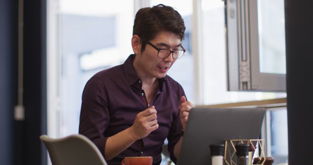 A businessman in an office setting is excitedly celebrating a success while working on his laptop. He is wearing glasses and a shirt, displaying a look of triumph. This image is ideal for illustrating concepts of business success, professional achievement, remote work, and motivation in a corporate environment. It can be used in business presentations, advertisements, websites, or articles about workplace success and professional development.