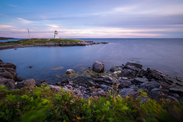 Sunset lighthouse scene with rocky shoreline is capturing a coastal landscape bathed in soft evening light. The serene setting creates a peaceful atmosphere, ideal for travel and tourism promotions, nature and maritime blogs, or as a relaxing wall art piece for homes and offices.